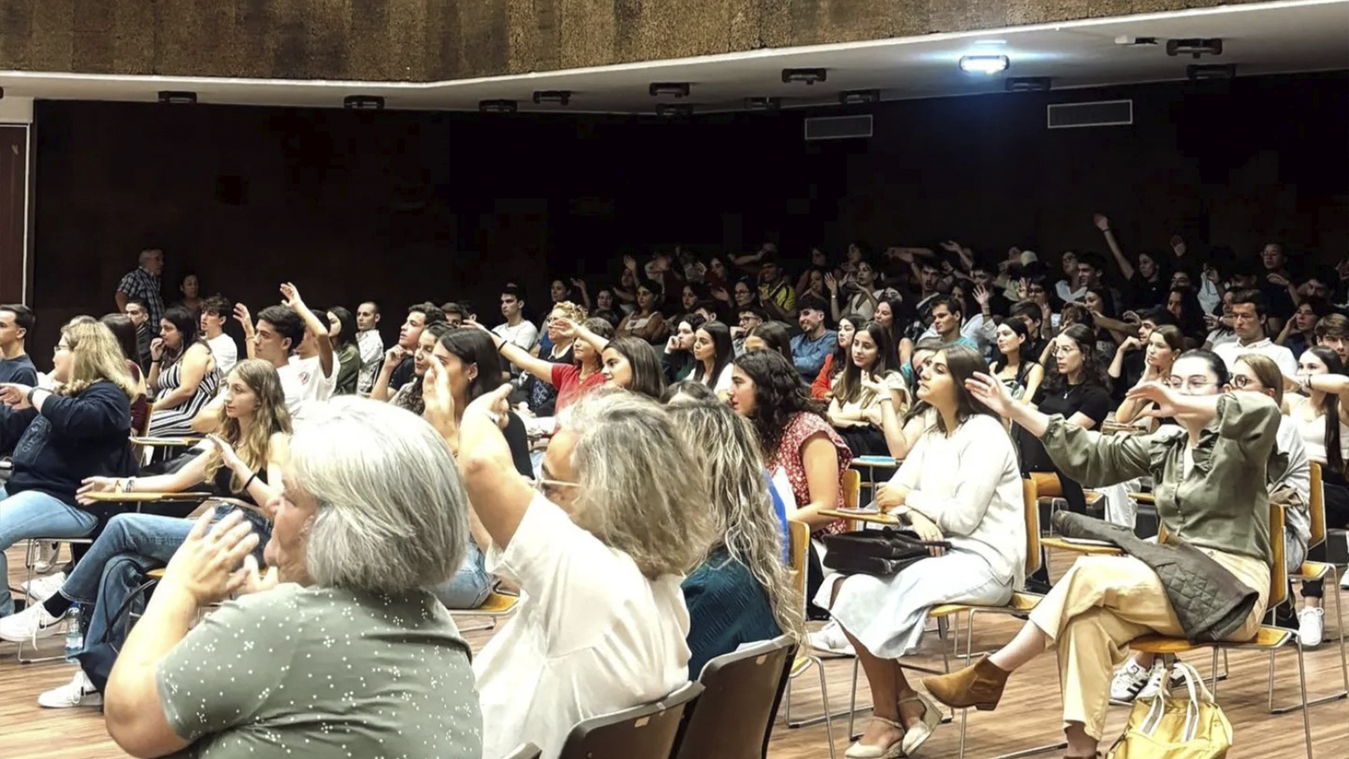 Grupo numeroso de pessoas em uma sala, todas com as mãos levantadas, demonstrando participação e engajamento.