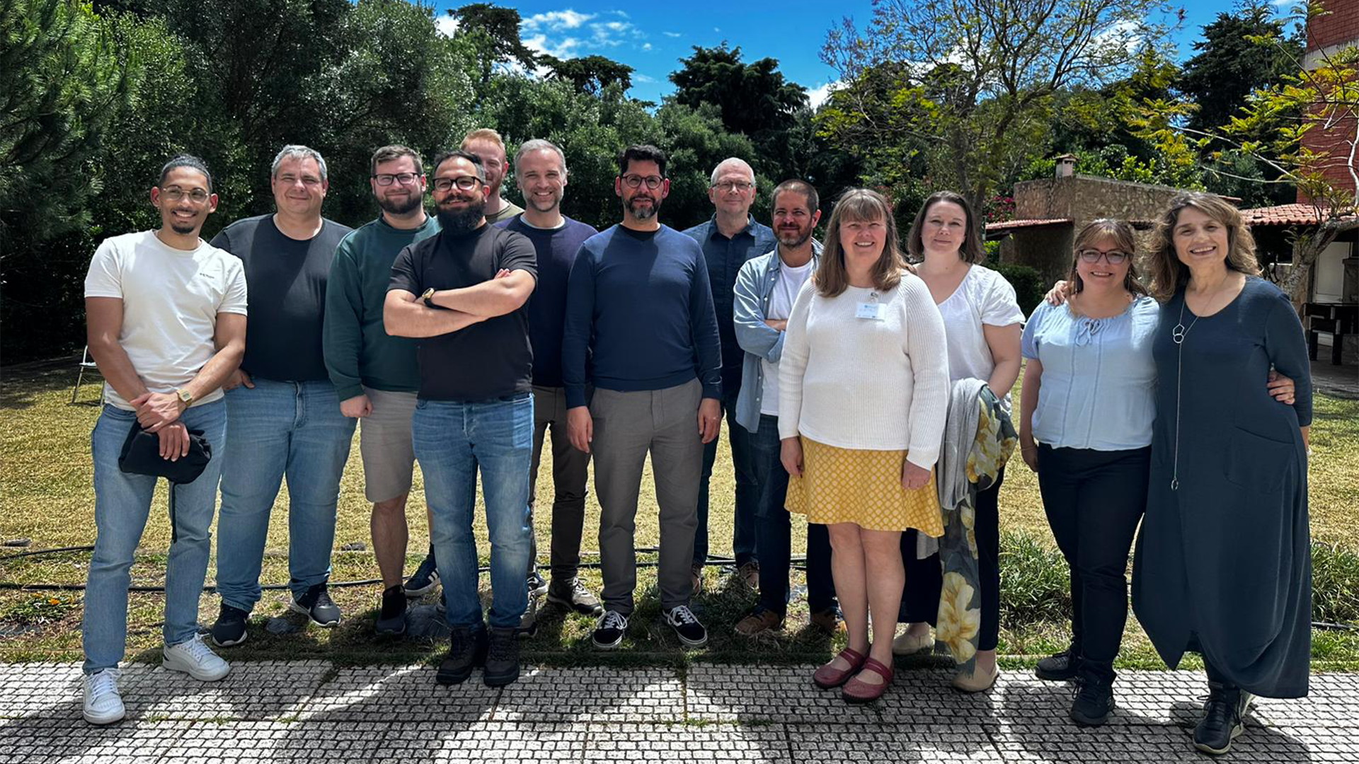 grupo de 14 pessoas posando ao ar livre em um dia ensolarado, com árvores ao fundo